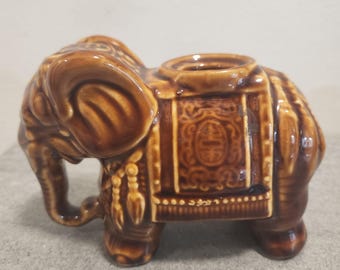 May include: A brown glazed ceramic elephant figurine featuring a decorative saddle with an embossed pattern and a circular opening on the back. The elephant is depicted in a standing position with its trunk curved downward and detailed textures.