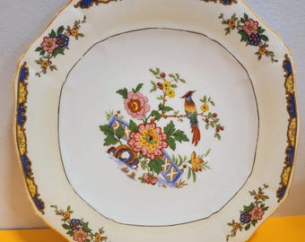 May include: Cream-colored octagonal plate with a floral and bird design. The center features a colorful bird perched on a branch with flowers. The rim has a blue and gold border with floral accents. A vintage decorative piece.