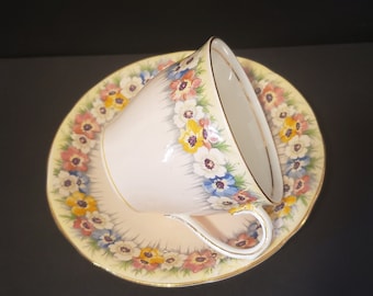 May include: A pink teacup and saucer with a floral pattern in shades of yellow, blue, pink, and green. The cup and saucer have a gold trim.