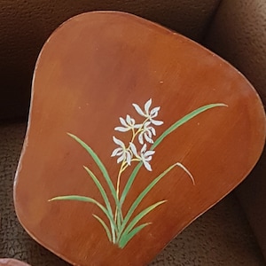 May include: A wooden coaster featuring a painted design of white flowers and green leaves. The coaster is brown and has a rounded shape.