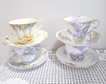 May include: Two stacks of porcelain teacups and saucers. The left stack features a cream cup with yellow flowers and a saucer with blue floral patterns. The right stack includes a white cup with purple bellflowers and a saucer with purple floral motifs.