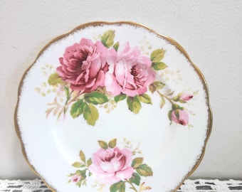 May include: A white ceramic plate with a scalloped edge and a gold-trimmed rim. The plate features a floral pattern with clusters of pink and deep rose-colored roses, green leaves, and small buds arranged in the center and along the lower section.