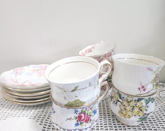 May include: A collection of porcelain teacups and saucers featuring various floral patterns. The cups display designs of pink roses, yellow blossoms, and white flowers, all accented with gold rims. The saucers are stacked, showing matching floral motifs.