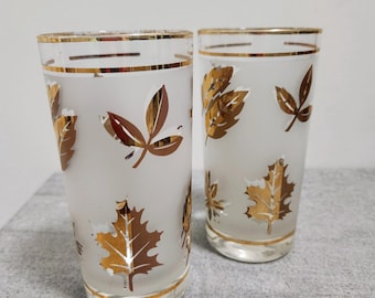 May include: Two vintage highball glasses with a frosted finish and gold leaf designs. The glasses have a gold rim and leaf patterns in shades of gold and brown. The glasses are likely from the mid-century era.
