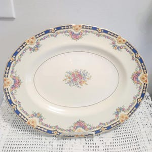 Peut inclure: Un plat de service ovale en céramique avec un centre de couleur crème orné d'un bouquet floral central. Le bord festonné est décoré d'une large bordure bleu marine, d'un liseré doré et d'un motif continu de petites fleurs roses, jaunes et bleues.