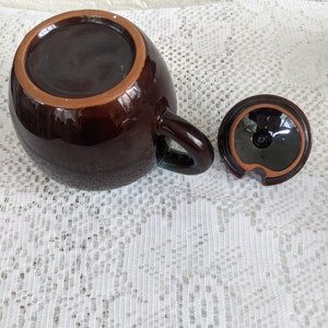 Op de afbeelding: Een bruine theepot van keramiek met een deksel en een handvat. De theepot staat op een wit kantkleed.