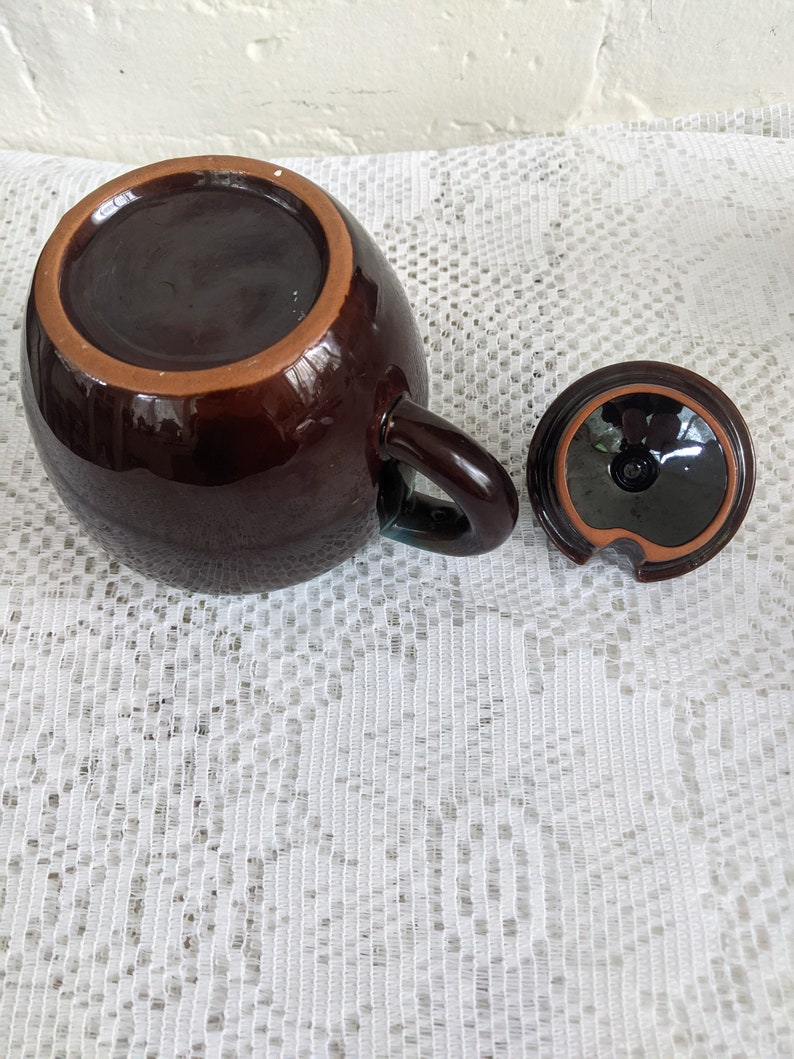 Op de afbeelding: Een bruine theepot van keramiek met een deksel en een handvat. De theepot staat op een wit kantkleed.