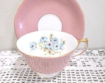 May include: A pink teacup and saucer set featuring a white interior with a blue floral bouquet design. The teacup has a gold-trimmed rim and handle, while the saucer features a matching pink surface with a gold-trimmed edge and white center.