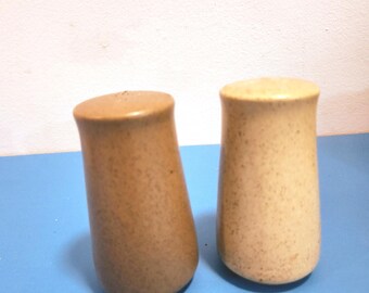 May include: Two ceramic salt and pepper shakers. The shakers are cylindrical with rounded tops and a speckled, earthy brown and tan color. The shakers are on a blue surface.