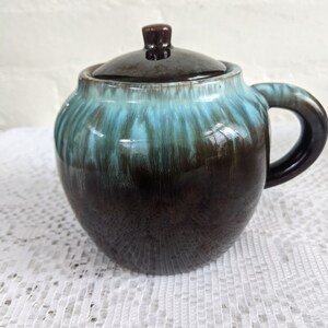 Op de afbeelding: Een bruine en teal geglazuurde keramische pot met een deksel en een handvat. De pot heeft een gemarmerde glazuur met strepen van teal en bruin.