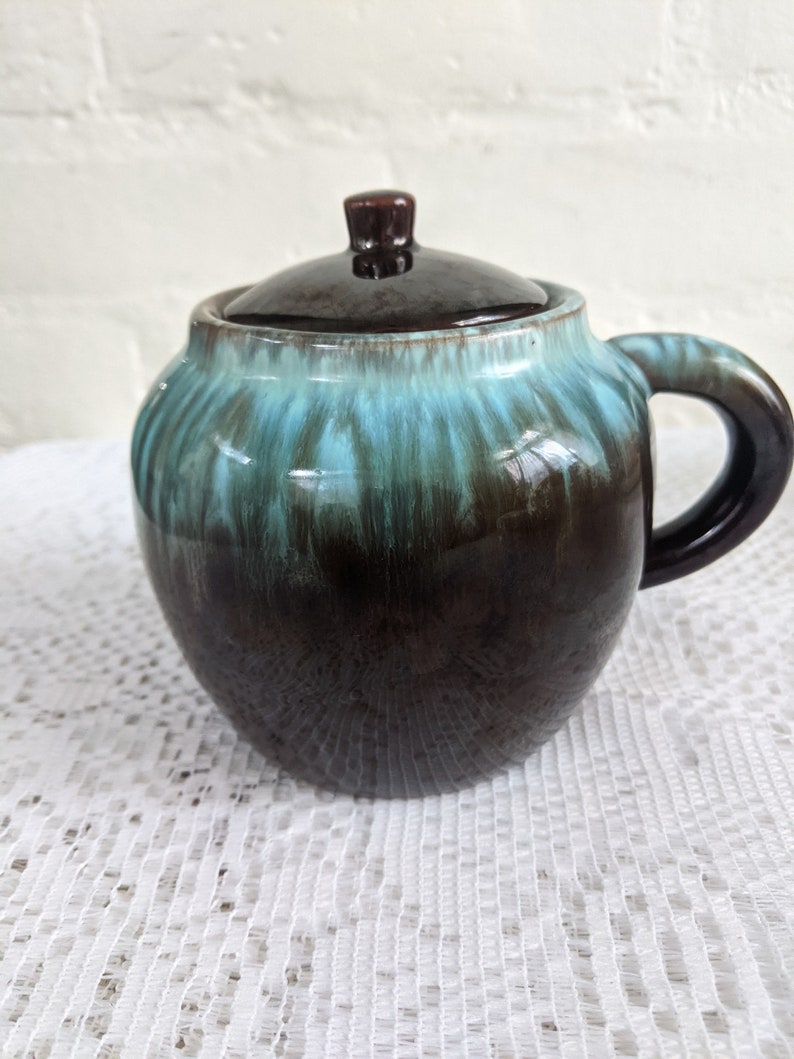 Op de afbeelding: Een bruine en teal geglazuurde keramische pot met een deksel en een handvat. De pot heeft een gemarmerde glazuur met strepen van teal en bruin.