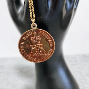 May include: A gold chain necklace with a round pendant featuring a gold coin with the inscription "II REGINA CORONAT II ELIZABETH II CANADA" and the royal cypher "EIIR".