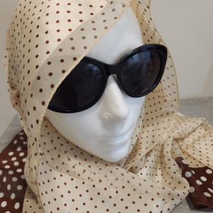 以下が含まれることがあります： 白いマネキンの頭に、茶色と白のドット柄のスカーフと黒い楕円形のサングラスをかけています。