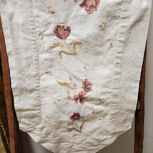 May include: A white linen table runner with embroidered pink and gold flowers. The runner has a scalloped edge and is perfect for adding a touch of elegance to any table setting.