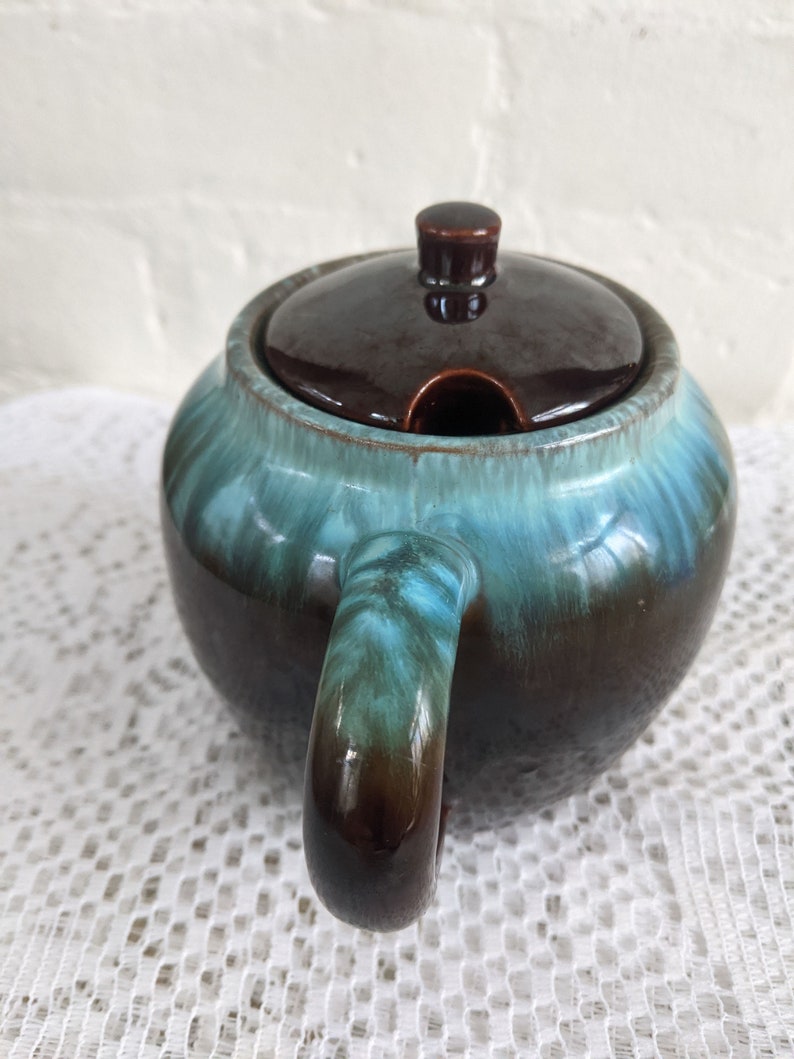 Op de afbeelding: Een bruine en blauwe geglazuurde keramische suikerpot met een deksel en een handvat. De kom heeft een gemarmerde glazuur met bruine en blauwe swirls.