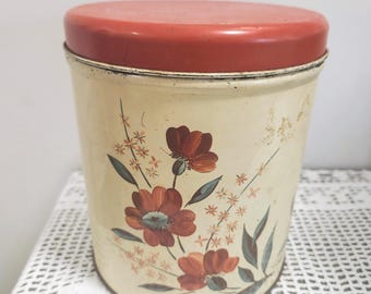 May include: A cylindrical metal storage canister with a cream-colored body and a solid red lid. The exterior features a painted floral design with dark red flowers, green leaves, and small yellow star-shaped accents on the front surface.