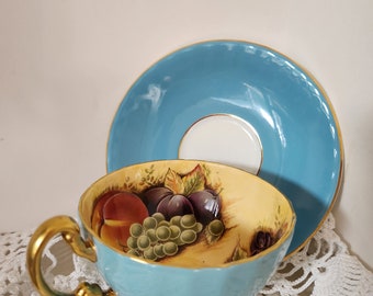 May include: A blue teacup and saucer with a gold rim. The teacup features a painted design of fruit, including grapes, plums, and peaches.