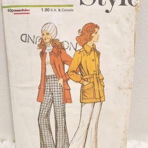 May include: Vintage sewing pattern labelled "Style" with illustrations of two women in 1970s attire. One wears a rust-coloured coat and plaid trousers, the other a yellow coat and white trousers. The pattern is for Miss size 10, bust 82.5 cm.