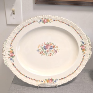 Peut inclure: Un plat de service ovale en céramique à fond crème, orné d'un bouquet floral central et d'une bordure décorative de petites fleurs colorées. Le plat présente un bord festonné et cannelé avec de subtiles touches vertes sur les poignées.