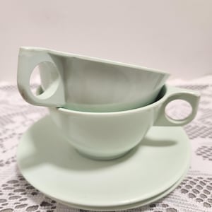 May include: Two light green teacups with matching saucers. The cups have a rounded bowl shape with a handle. The saucers are round and flat. The teacups are stacked on top of each other, with one resting on a saucer.