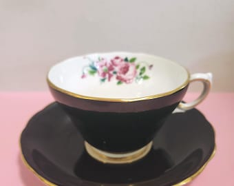 May include: A black teacup and saucer set with gold trim. The inside of the teacup is white with a floral design of pink roses. The saucer is round and has a matching gold trim. The teacup has a handle.