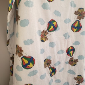 May include: White fabric featuring a repeating pattern of colourful hot air balloons and teddy bears. The balloons are red, yellow, green, and blue. Light blue clouds are scattered across the background.