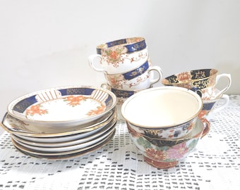 May include: A collection of porcelain teacups and saucers featuring a white base with navy blue and gold trim, accented by orange and pink floral patterns. The set includes stacked saucers and multiple teacups with curved handles and gold rims.