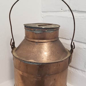 Peut inclure: Une cruche à lait en cuivre avec une poignée en fil de fer et un couvercle. La cruche a un aspect usé et vieilli.