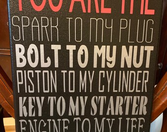 May include: Black canvas sign with white and gray text that reads "You are the spark to my plug, bolt to my nut, piston to my cylinder, key to my starter, engine to my life. I love you."  The sign is decorated with two gray metal gears on either side of the text "I love you".