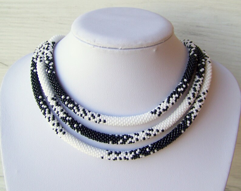 May include: A black and white beaded necklace with a multi-strand design. The necklace is made of small, round beads that are woven together in a pattern.