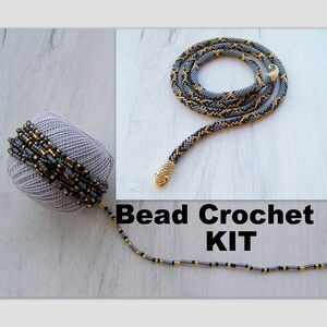 Może przedstawiać: Zestaw do robienia na szydełku z koralików do stworzenia naszyjnika w kształcie węża. Zestaw zawiera kłębek koralików w kolorze szarym i złotym, złoty wisiorek w kształcie głowy węża i nić do koralików. Gotowy naszyjnik jest pokazany na zdjęciu.