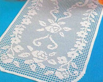 May include: White crocheted table runner with a floral design. The runner is on a blue tablecloth.