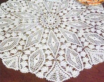 Può includere: Un tovaglia rotonda in pizzo bianco all'uncinetto con un delicato motivo floreale si trova su un tavolo di legno. La tovaglia ha un diametro di circa 46 cm.