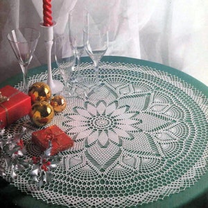 Puede incluir: Un mantel de mesa blanco de ganchillo con un delicado diseño floral, colocado sobre un mantel verde. El mantel está rodeado de vasos, un candelero y adornos decorativos.