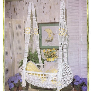 Op de afbeelding: Een witte macramé babywieg met een geel en wit kussen, hangend aan een touw. De wieg is omgeven door paarse bloemen in potten en een schilderij van een zeilboot. De tekst "Juliano's Hang It All" staat bovenaan.