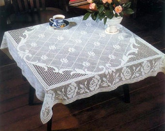 May include: White crocheted tablecloth with a floral design, featuring a delicate pattern of roses and leaves. The tablecloth is laid on a wooden table with a cup and saucer on top.
