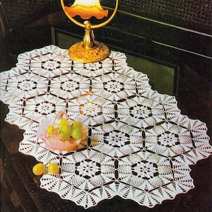 May include: White crocheted table runner with a delicate floral pattern. The runner is laid on a dark surface with a decorative lamp and a bowl of green grapes.