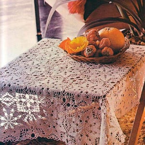 May include: A white crocheted lace tablecloth with an intricate floral pattern draped over a square table. A small woven basket sits on top, filled with fresh strawberries, a single orange, and a bright yellow poppy flower blossom.