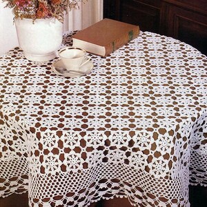 May include: A white, round, crocheted tablecloth with a detailed floral pattern. The tablecloth is draped over a table, with a teacup and saucer, a book, and a vase of flowers visible on top. The crochet work is intricate.
