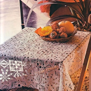 May include: A rectangular, off-white, crocheted lace tablecloth covers a table. A woven basket holds an orange, several small pumpkins, and a yellow flower. A bench with a colorful striped blanket is in the background.