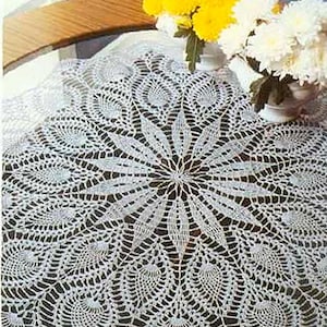 May include: A white, finely crocheted tablecloth with a central floral pattern. The delicate lace design features radiating lines and circular motifs. Yellow and white flowers in vases sit on the table, adding colour to the scene.