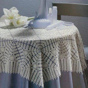 May include: A round, off-white crocheted tablecloth with an intricate, repeating floral lace pattern and a scalloped edge. The fabric features a delicate, open-work design that drapes over the sides of a circular table, showcasing fine thread work.