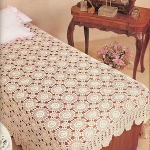 Könnte beinhalten: Eine cremefarbene, gehäkelte Tagesdecke mit einem sich wiederholenden kreisförmigen Muster bedeckt ein Bett. Ein passendes Kissen mit gerüschtem Rand liegt auf dem Bett. Ein hölzerner Schminktisch mit Spiegel, Lampe und Parfümflaschen steht daneben. Ein Strauß rosa Blumen befindet sich auf dem Schminktisch.