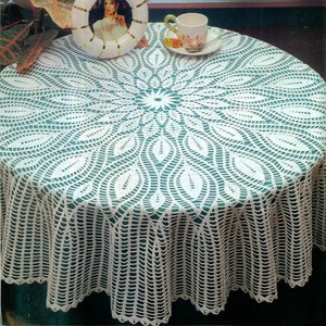 Peut inclure: Nappe en crochet blanc avec un motif floral délicat. La nappe est posée sur une table ronde avec une surface verte. Un cadre photo et une tasse et une soucoupe sont sur la table.