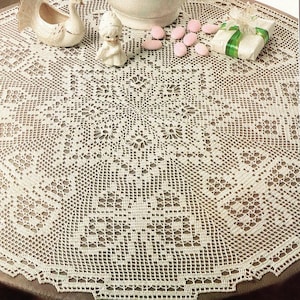 Puede incluir: Un mantel de mesa de crochet blanco con un delicado patrón floral. El mantel está puesto sobre una mesa con un jarrón blanco lleno de flores rosas, un pájaro de porcelana blanca y un pequeño ángel de porcelana blanca.