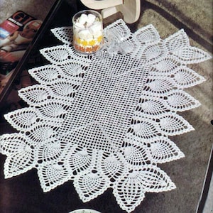Op de afbeelding: Een witte gehaakte tafelloper met een delicaat, kantpatroon met een herhalend bladmotief. De loper ligt op een glazen tafelblad.