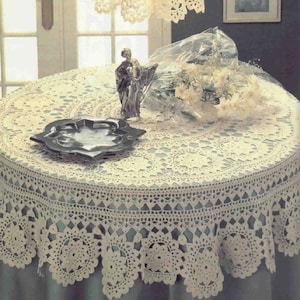 May include: A white crocheted tablecloth with a delicate lace pattern. The tablecloth is round and has a scalloped edge. It is laid on a table with a silver plate and a decorative figurine.