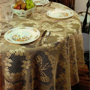 May include: A crocheted tablecloth with a delicate floral pattern in a cream colour. The tablecloth is laid on a wooden table with a bowl of fruit, glasses, and plates.
