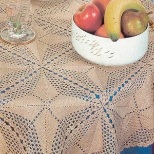 May include: A beige crocheted tablecloth with a geometric pattern. A white bowl filled with fruit, including a red apple, orange, banana, and pear, sits on the table. A glass of water and a coaster are also on the table.