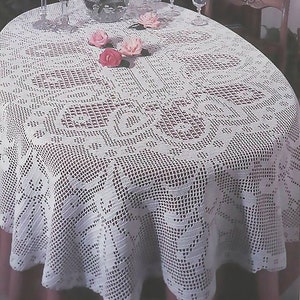 Może przedstawiać: Biały owalny obrus szydełkowy z delikatnym wzorem kwiatowym. Obrus leży na stole z różowym obrusem pod spodem. Na stole stoi wazon z różowymi różami.